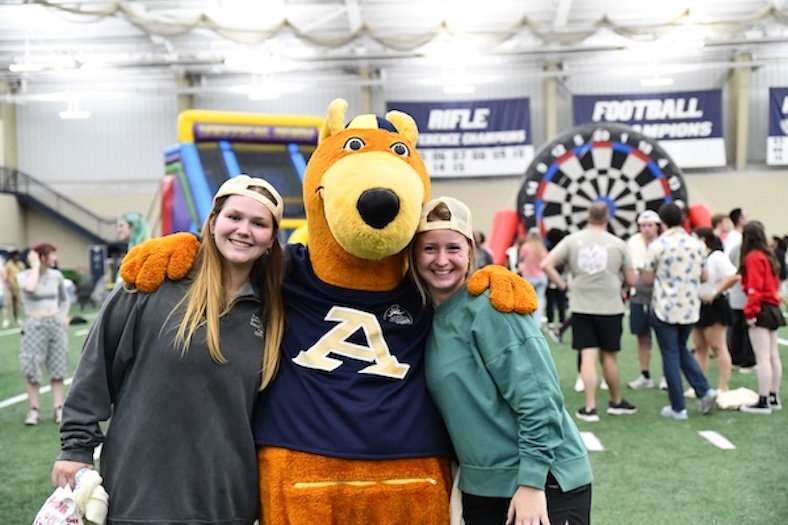Two smiling people in backwards caps pose with a kangaroo mascot wearing a blue shirt with an A on it, inside a gym with games and other people in the background.