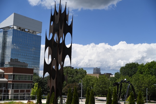 A tall, abstract metal sculpture with circular cutouts stands in front of modern glass and brick buildings, with trees and a partly cloudy sky in the background.