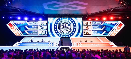 A live esports event features two teams, UNT and Akron, competing on stage with large digital screens showing player names and scores. An audience watches the match under colorful lighting and a prominent event logo.