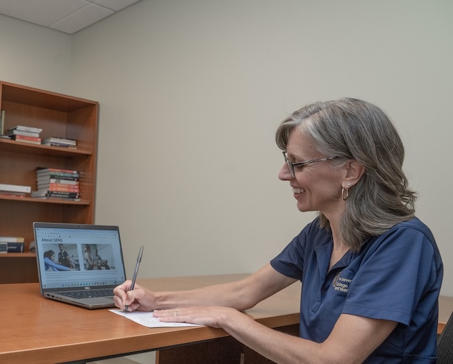 A woman with gray hair and glasses writes on paper at a desk while smiling at an open laptop displaying a website. Books are stacked on shelves in the background.