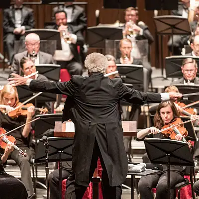 A conductor leads an orchestra in performance, with musicians playing violins, cellos, and brass instruments on stage.