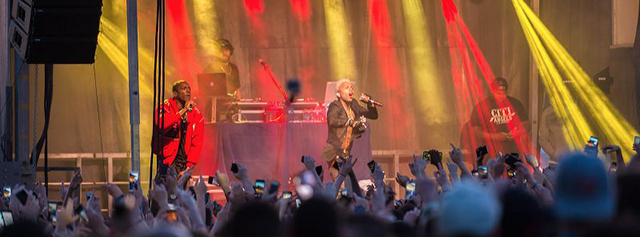 A large crowd at an outdoor concert raises their hands and phones as two performers sing and rap on stage with bright yellow and red lights behind them. A DJ and another person stand near the back of the stage.