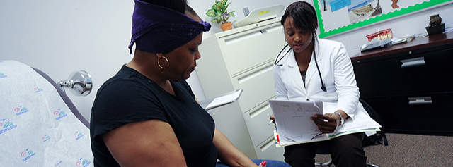A doctor in a white coat reviews paperwork while speaking to a patient sitting on an exam table in a medical office. The patient looks down, and the room has medical equipment and a file cabinet in the background.