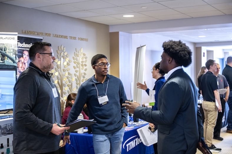 Three men talk at a career fair, with two holding clipboards. Booths with banners and other attendees are visible in the background.