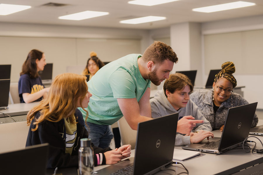 A man leans over to help two students with laptops in a classroom, while another student smiles and looks on. Other students work in the background.