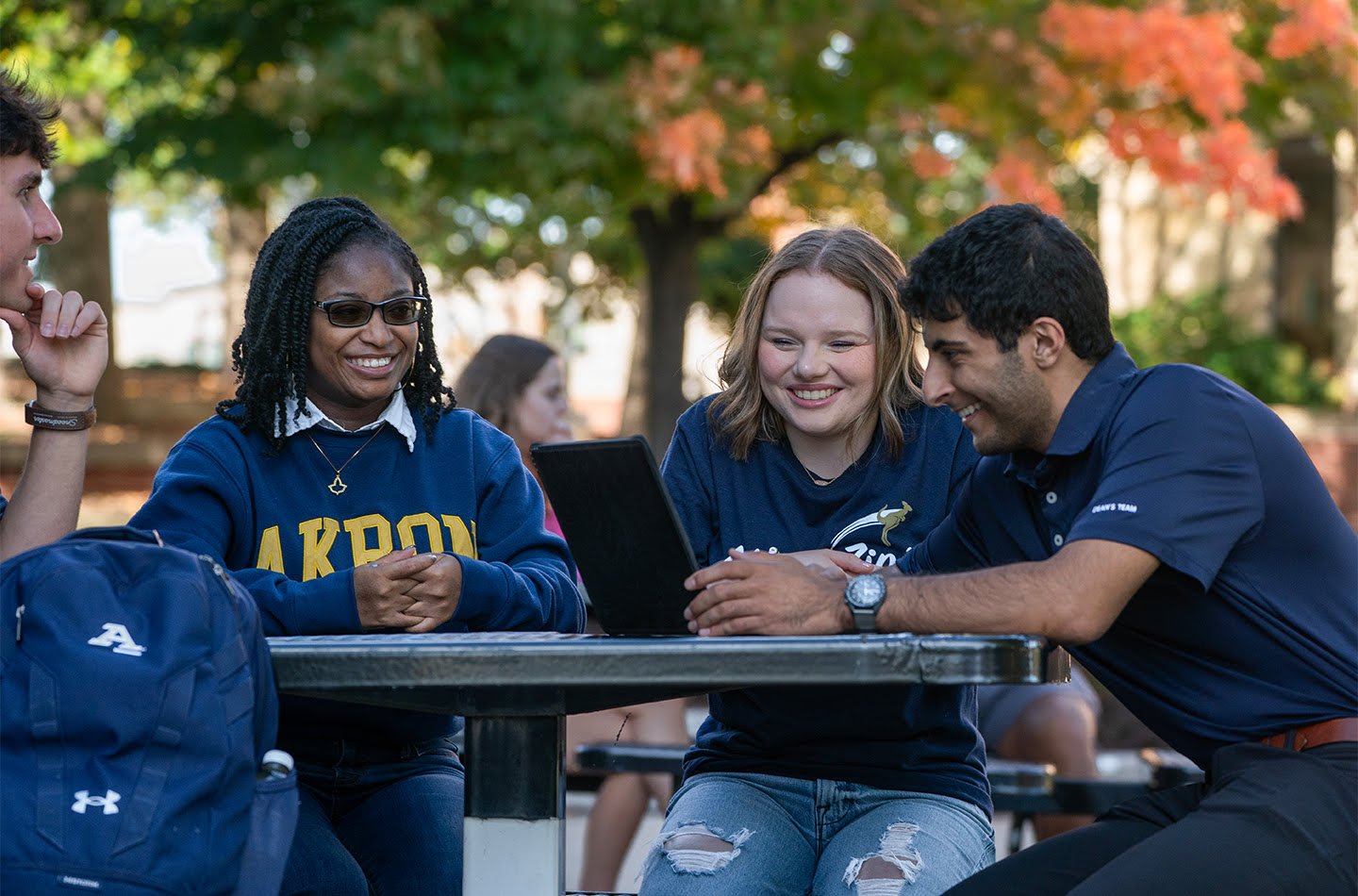 A group of students sitting outside at a picnic table, having a good time, while one student shows them some work on a laptop