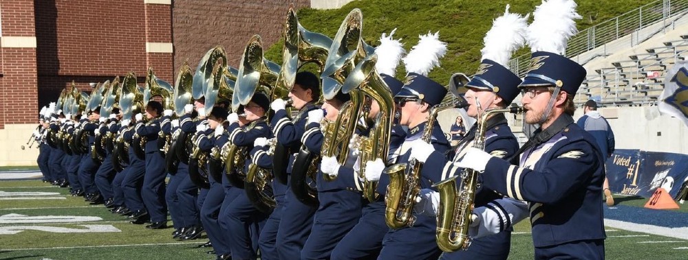 A marching band in blue uniforms and feathered hats performs on a football field, playing brass instruments in a synchronized row. Stadium seating and a brick wall are visible in the background.