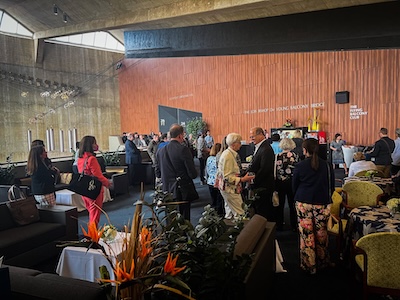 A group of people in formal attire mingle and converse in a modern lounge with wooden walls, large windows, and contemporary furniture. Bright flowers decorate the tables.