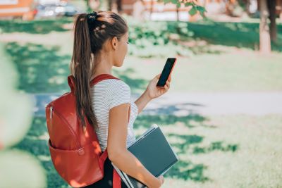 A side-profile view of a student walking outdoors on a sunny campus. She has her hair in a high ponytail, wears a red backpack, and holds notebooks under her arm while looking down at a smartphone in her hand.
