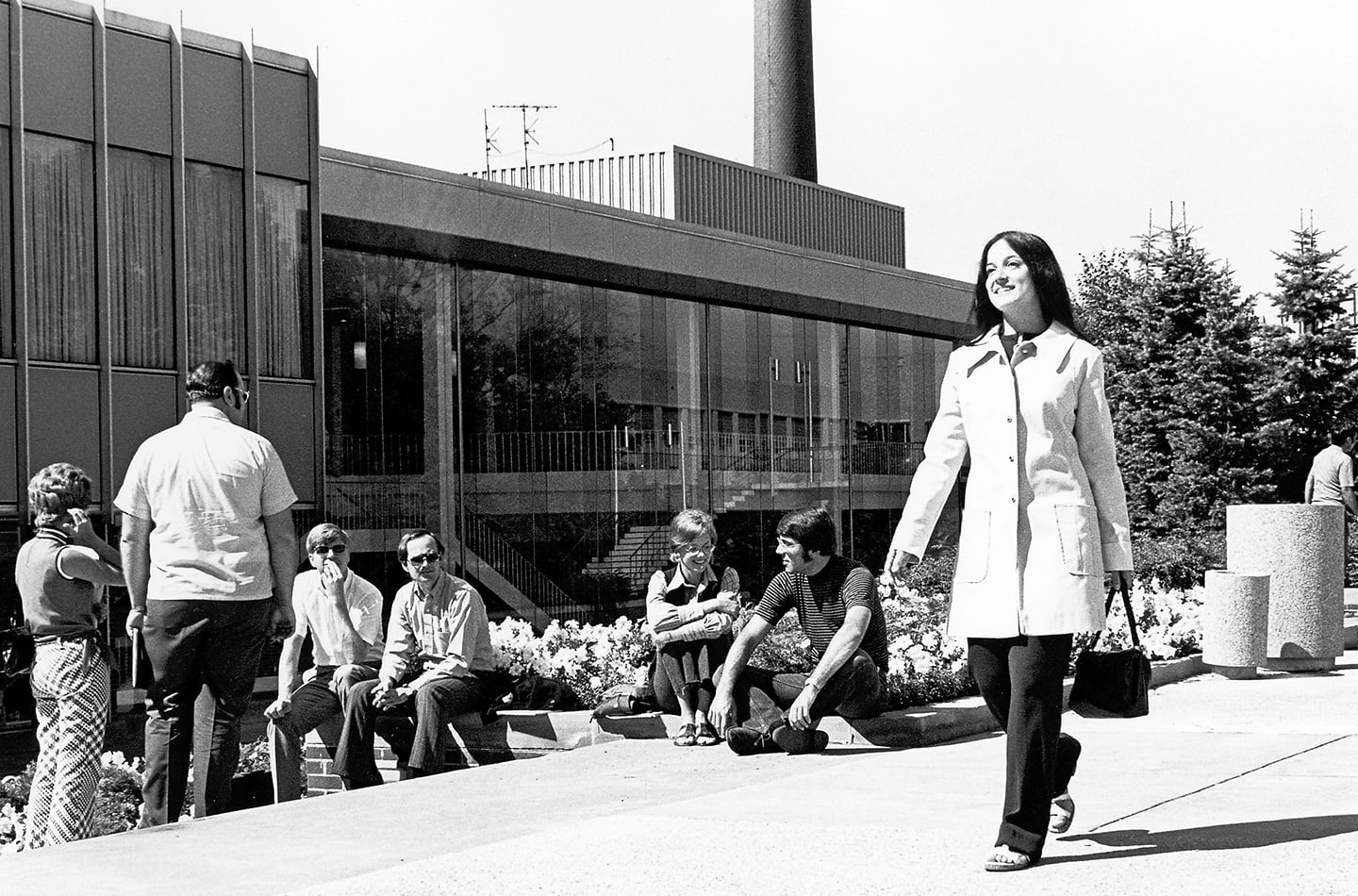 A black and white historical photo of a woman in a 1960s-style trench coat walking on a university campus, with several other students sitting on a low wall in front of a modern glass-walled building in the background.