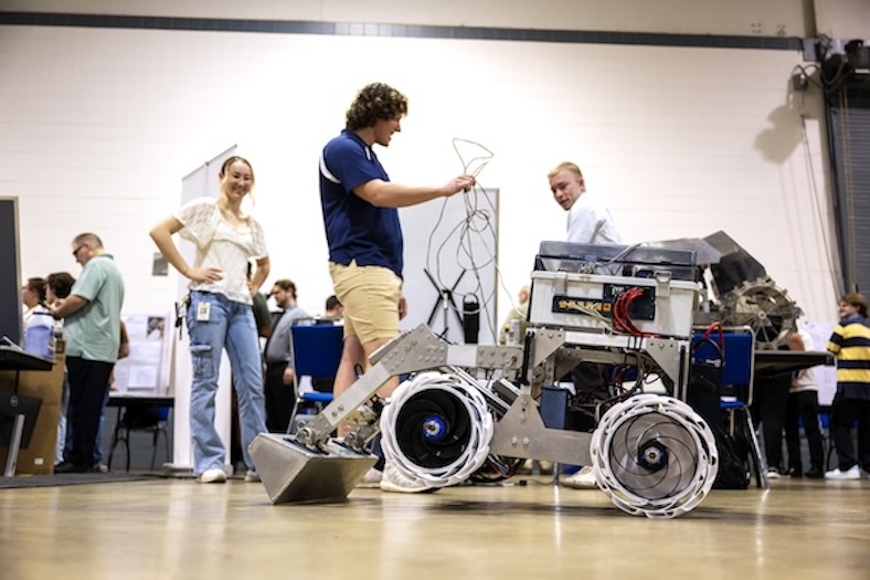 Students interact with a wheeled robot at an indoor event. One student holds a controller while others watch and smile. People and display tables are visible in the background.
