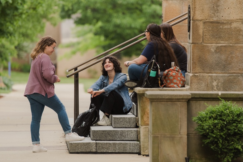 Four young women are talking and laughing together outside on stone steps near a building, with backpacks and water bottles beside them. Trees and greenery are visible in the background.