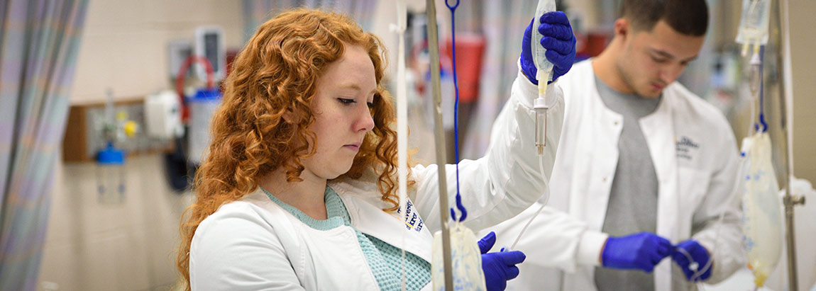 Two healthcare professionals in white coats and purple gloves prepare IV bags in a clinical setting, with medical equipment visible in the background.