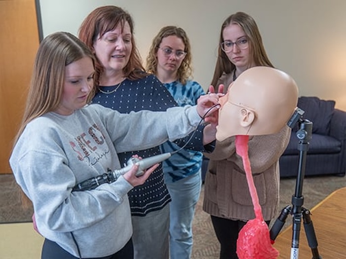 Four women observe as one uses a medical instrument on a human head and neck model mounted on a stand, practicing a procedure together in an indoor educational setting.