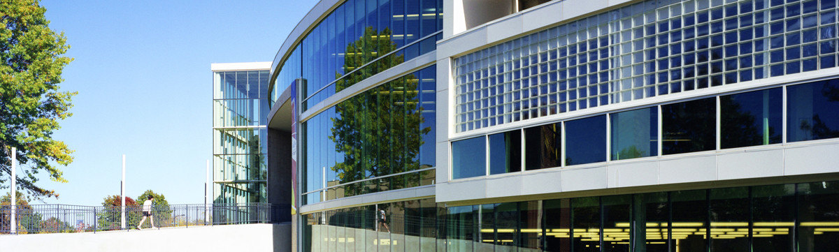 A wide exterior shot of a modern, multi-story academic or office building featuring a curved glass facade and white paneling. The large glass windows reflect the surrounding green trees and a clear blue sky.