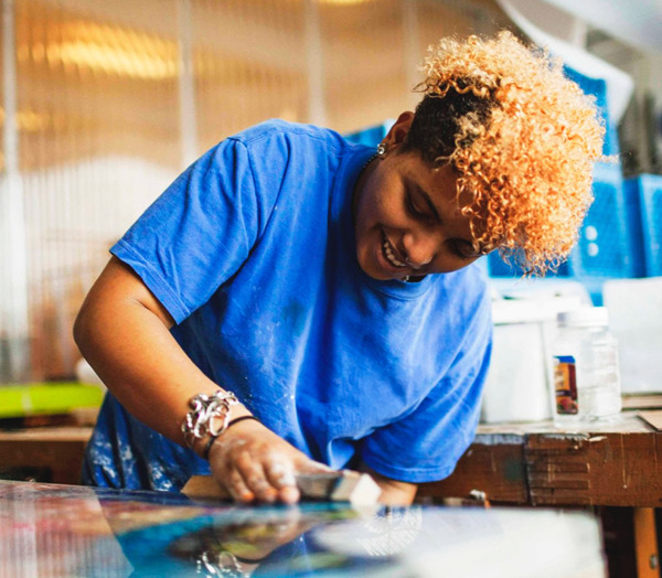A person with curly, dyed blonde hair wearing a blue shirt smiles while working with a tool on a flat surface in an art studio or workshop.