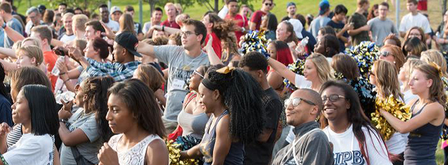 A large, diverse group of students gathers outdoors, cheering and smiling. Some wear matching shirts and hold pom-poms, suggesting a pep rally or school event. The atmosphere is lively and energetic.