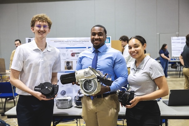 Three people stand in front of a project display holding mechanical devices, smiling at the camera. Poster boards and other people are visible in the background of the indoor setting.