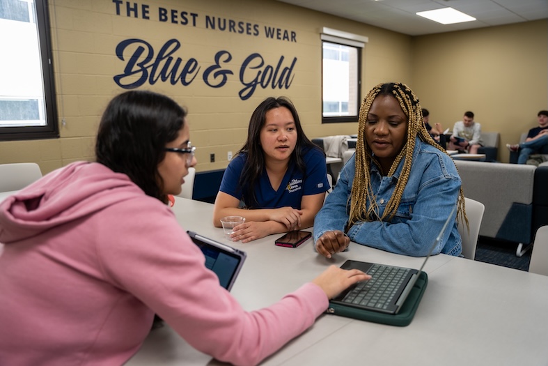 Three students sit at a table, working together with a laptop. A wall behind them says, The Best Nurses Wear Blue & Gold. Other students are seated in the background.