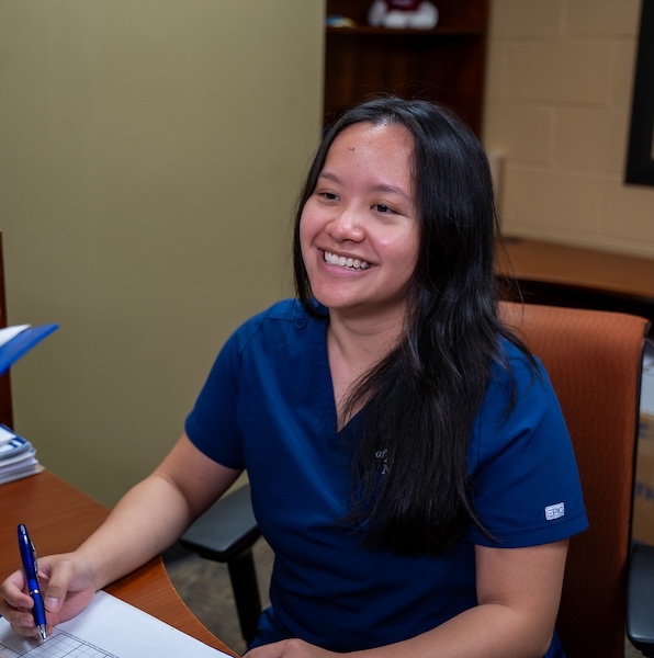 A smiling woman in blue scrubs sits at a desk, holding a pen and writing on a document. She appears to be in an office or medical setting with shelves and papers in the background.