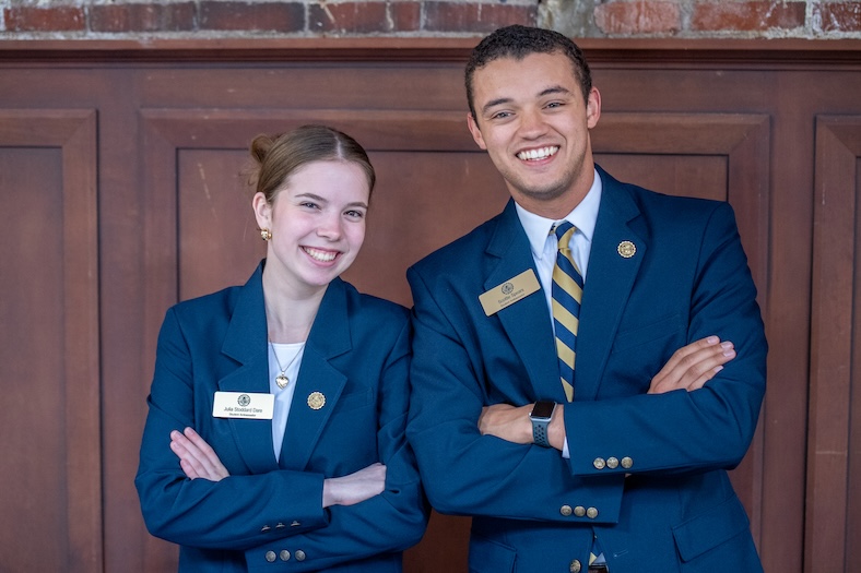 Two people wearing blue blazers, name tags, and pins stand side by side with arms crossed, smiling in front of a wooden wall with a brick detail above.
