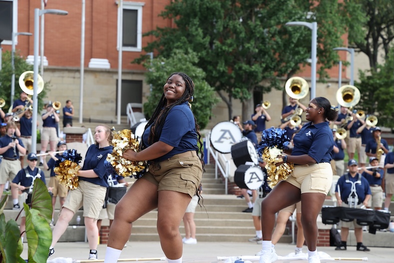 A wide shot of student performers on an outdoor campus plaza. In the foreground, three dancers in navy polo shirts and khaki shorts perform a routine with gold and navy blue pom-poms. Behind them, members of the marching band are arranged on steps, playing instruments including tubas, saxophones, and drums. The band members wear matching navy polos and khaki shorts. A large brick university building and green trees are visible in the background.
