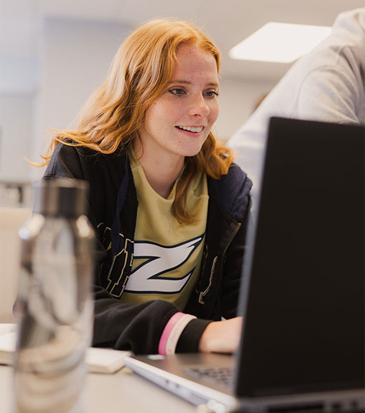 University of Akron student works on a laptop during class while wearing an Akron Zips T-shirt and hoodie.