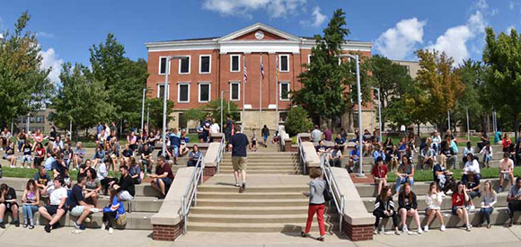 Large group of students sitting and gathering on wide outdoor steps and a plaza, with a red brick academic building and trees in the background under a blue sky with scattered clouds.