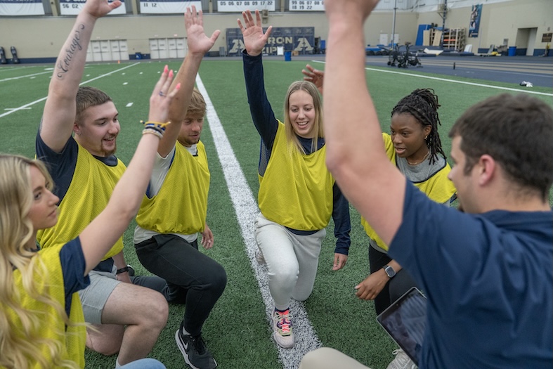 Six young adults wearing yellow pinnies kneel in a huddle on an indoor turf field, smiling and raising their hands together in a team cheer. A coach kneels nearby, joining in the gesture.