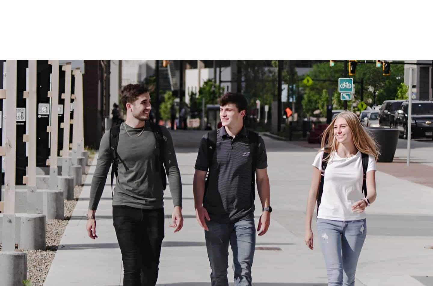 Three students walking and smiling together on a sunny city sidewalk in downtown Akron. In the background, the historic Akron Civic Theatre marquee is visible, while the foreground features several outdoor informational kiosks with QR codes.