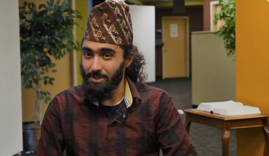 A man with a patterned cap and a beard sits indoors, wearing a maroon shirt. There is a table with an open book and potted plants in the background.