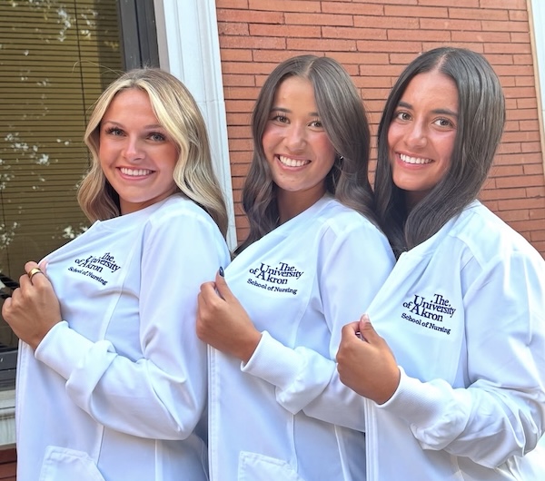 Three women smiling and standing side by side, wearing white lab coats with The University of Kentucky School of Nursing logo, posing in front of a red brick building.