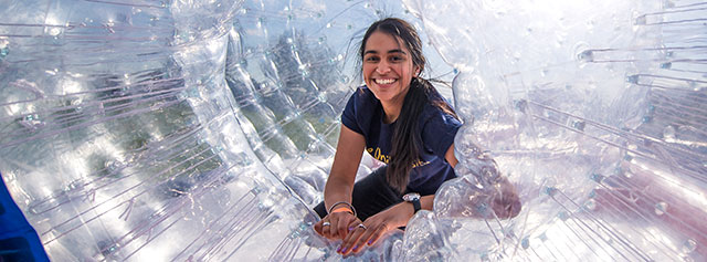 A young woman smiles brightly while inside a large, transparent inflatable ball, known as a zorb, with sunlight shining through the plastic.
