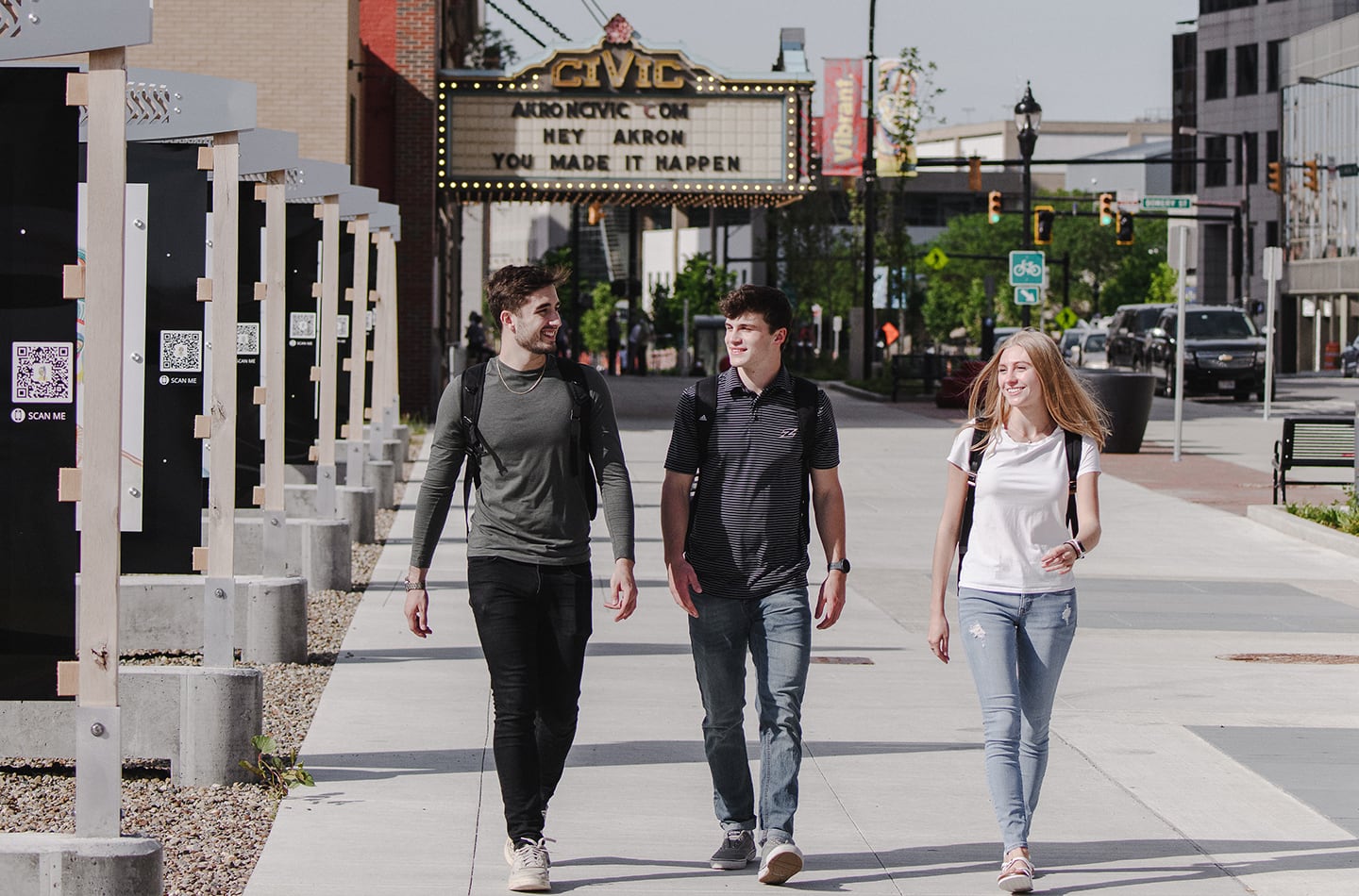 Three students walking and smiling together on a sunny city sidewalk in downtown Akron. In the background, the historic Akron Civic Theatre marquee is visible, while the foreground features several outdoor informational kiosks with QR codes.