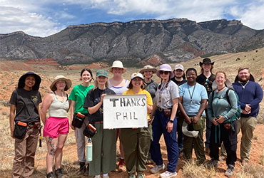 University of Akron geology field camp students stand together in a mountain landscape holding a sign that reads, 