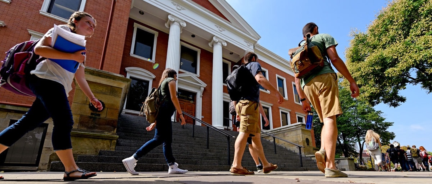 Students with backpacks walk past a red-brick academic building with white columns and stone steps on a sunny day.