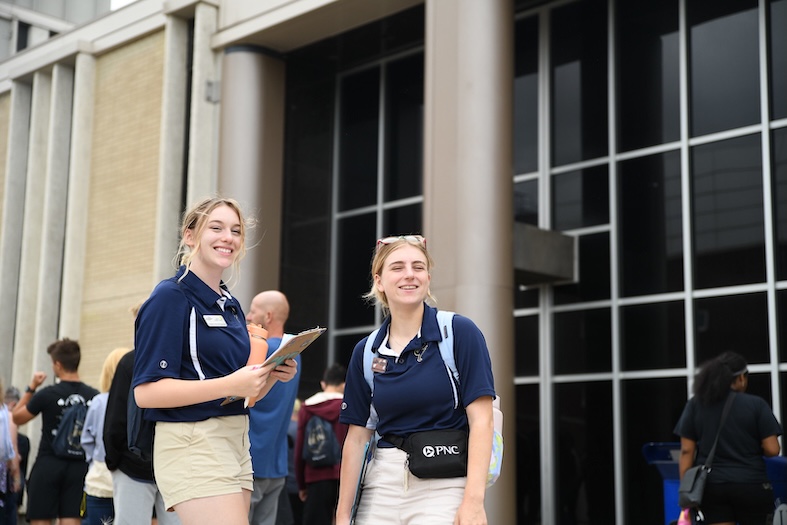 Two young women wearing navy shirts and khaki shorts stand smiling outside a building with tall windows and columns, holding a clipboard and a fanny pack, while people walk in the background.