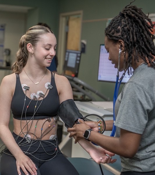 A student has her blood pressure measured by a classmate in an Exercise Science lab. She is seated and smiling, with ECG electrodes attached to her chest and torso connected to monitoring equipment.