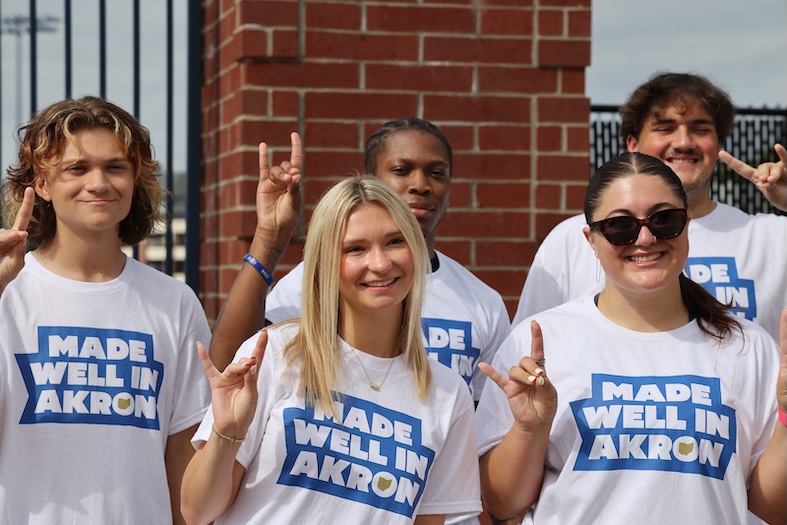 Five people wearing Made Well in Akron t-shirts smile and hold up their index fingers, posing outdoors in front of a brick structure.