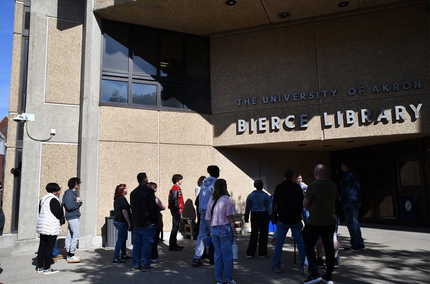 A group of students and visitors gathered outside the concrete entrance of the University of Akron Bierce Library on a sunny day.