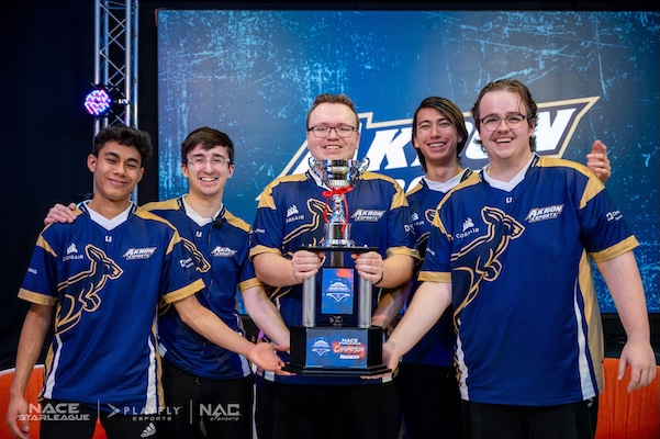 Five esports team members in matching blue jerseys smile and pose together on stage, holding a trophy. A logo is displayed on a screen behind them. Stage lighting and sponsor logos are visible in the background.