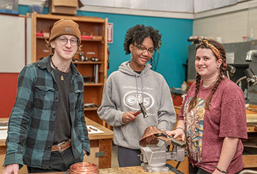 Three students in a workshop use tools to shape metal while working on a hands-on metalsmithing project.