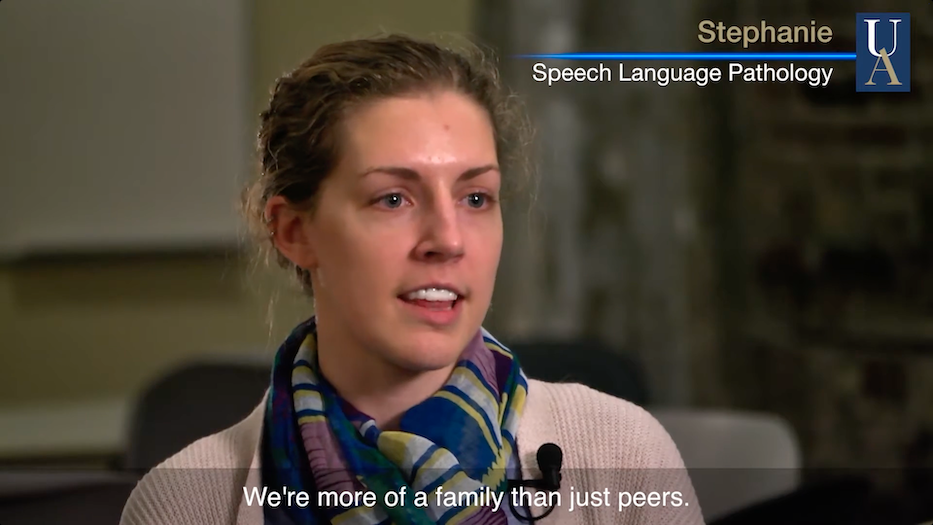 A woman with light brown hair pulled back, wearing a scarf and sweater, speaks indoors. The text reads: Stephanie, Speech Language Pathology and Were more of a family than just peers.
