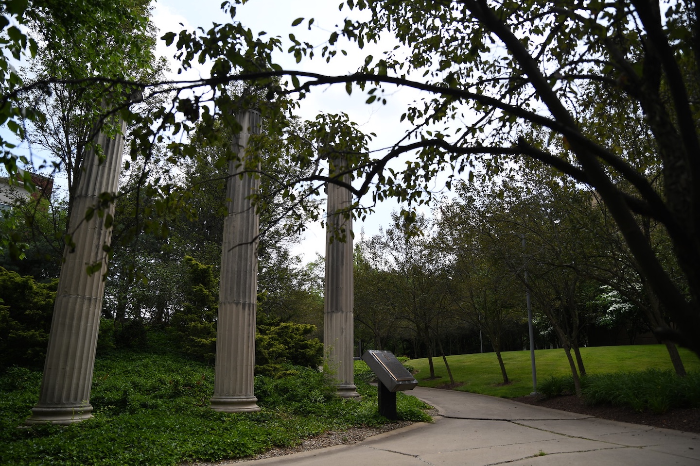 Four tall stone columns stand beside a paved walkway in a green park, surrounded by trees and grass, with an informational sign nearby.