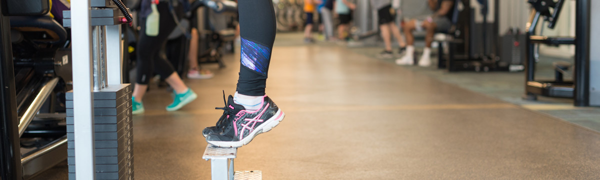 A close-up of a student's foot on a weight machine in a busy campus fitness center.