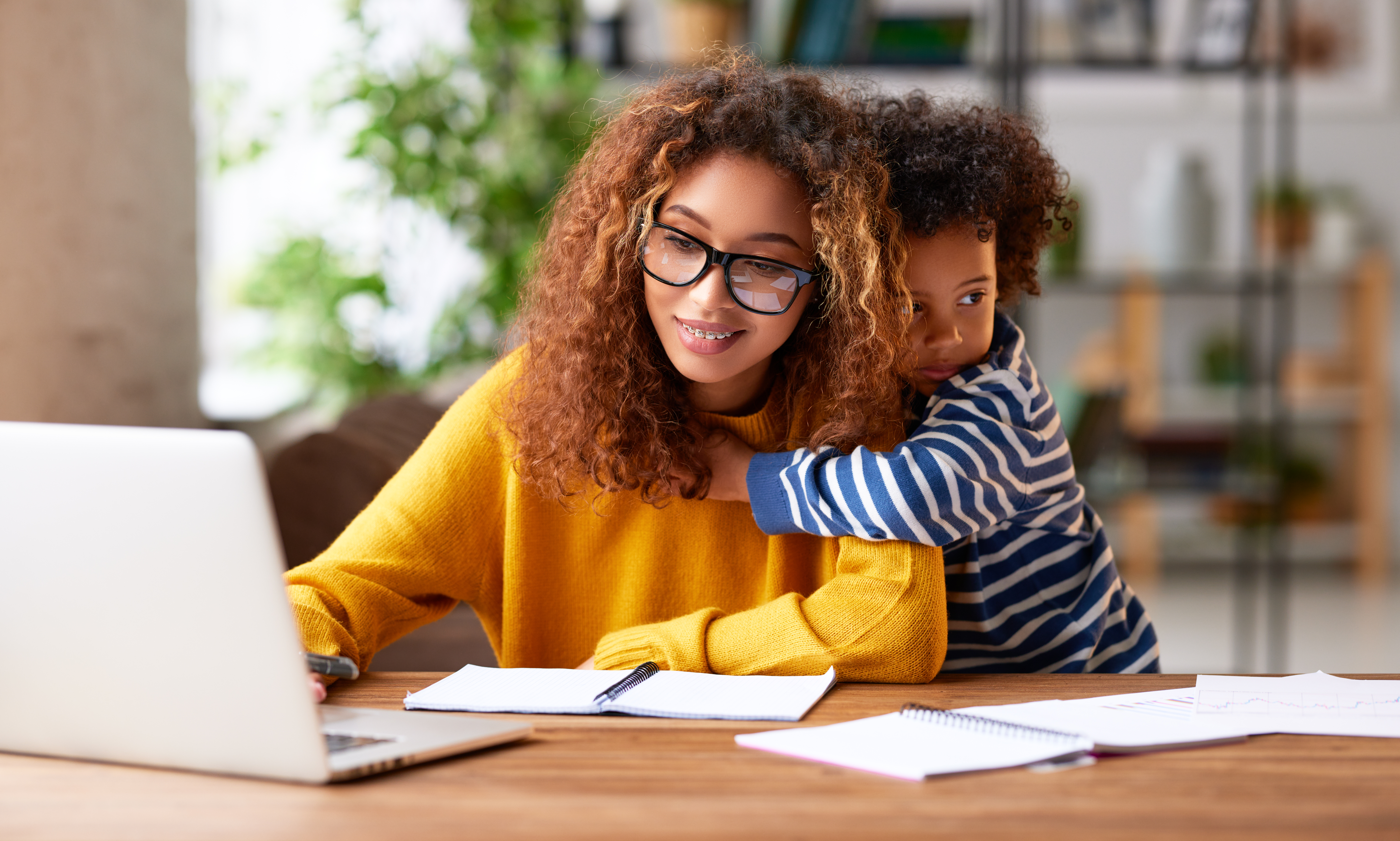 A woman in a yellow shirt working on a laptop while a young child in a blue and white striped shirt puts his arm around her.