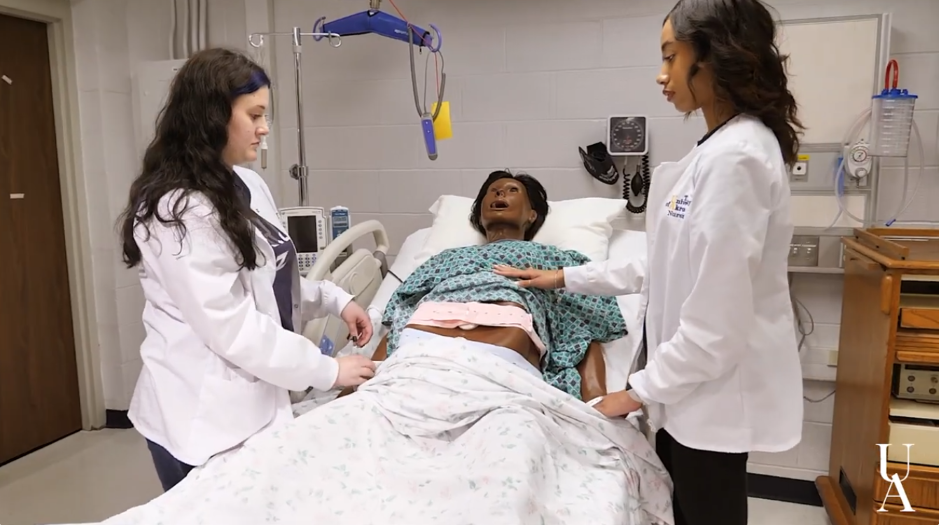 Two medical students in lab coats practice an abdominal exam on a medical mannequin in a hospital room simulation.