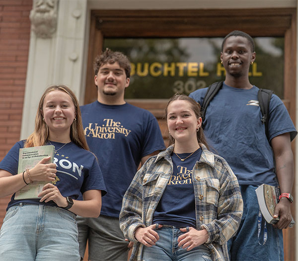 University of Akron students stand on the steps of Buchtel Hall holding books and smiling at the camera.
