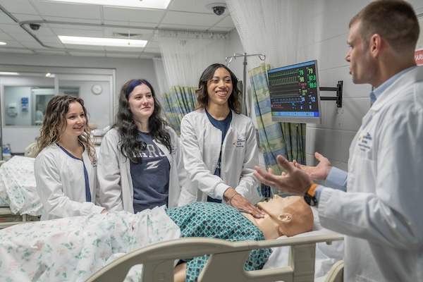 Three nursing students and an instructor in white lab coats are gathered around a high-fidelity medical simulation manikin lying in a hospital bed. The instructor, on the right, is speaking and gesturing toward the manikin, which is partially uncovered to show internal electronic components in the chest area.