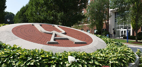 A large red and white UA logo is set in a circular brick area surrounded by green plants; a building entrance is visible in the background.